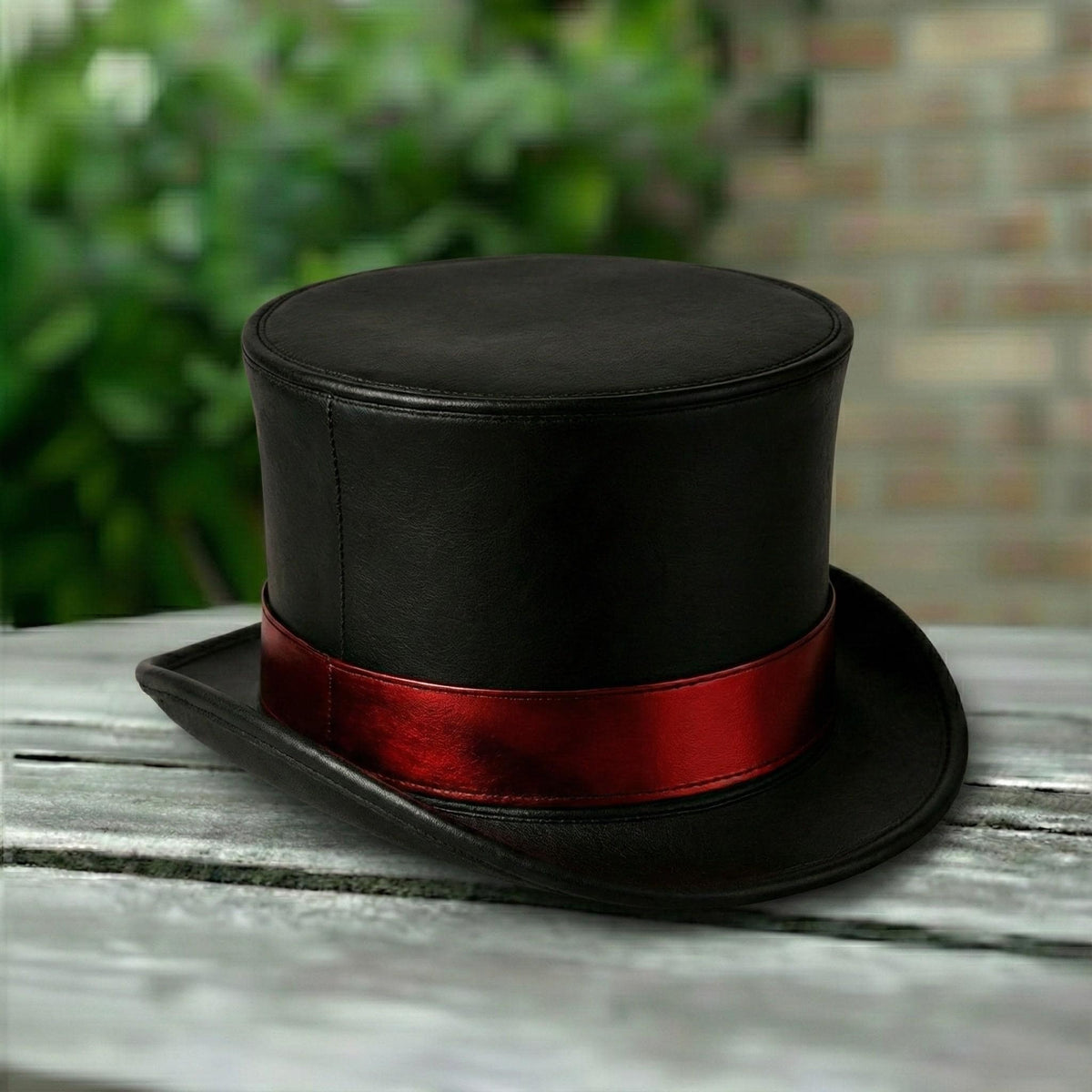 May include: A black top hat with a red band. The hat is made of a smooth material and sits on a wooden surface. The background features blurred green foliage and a brick wall.